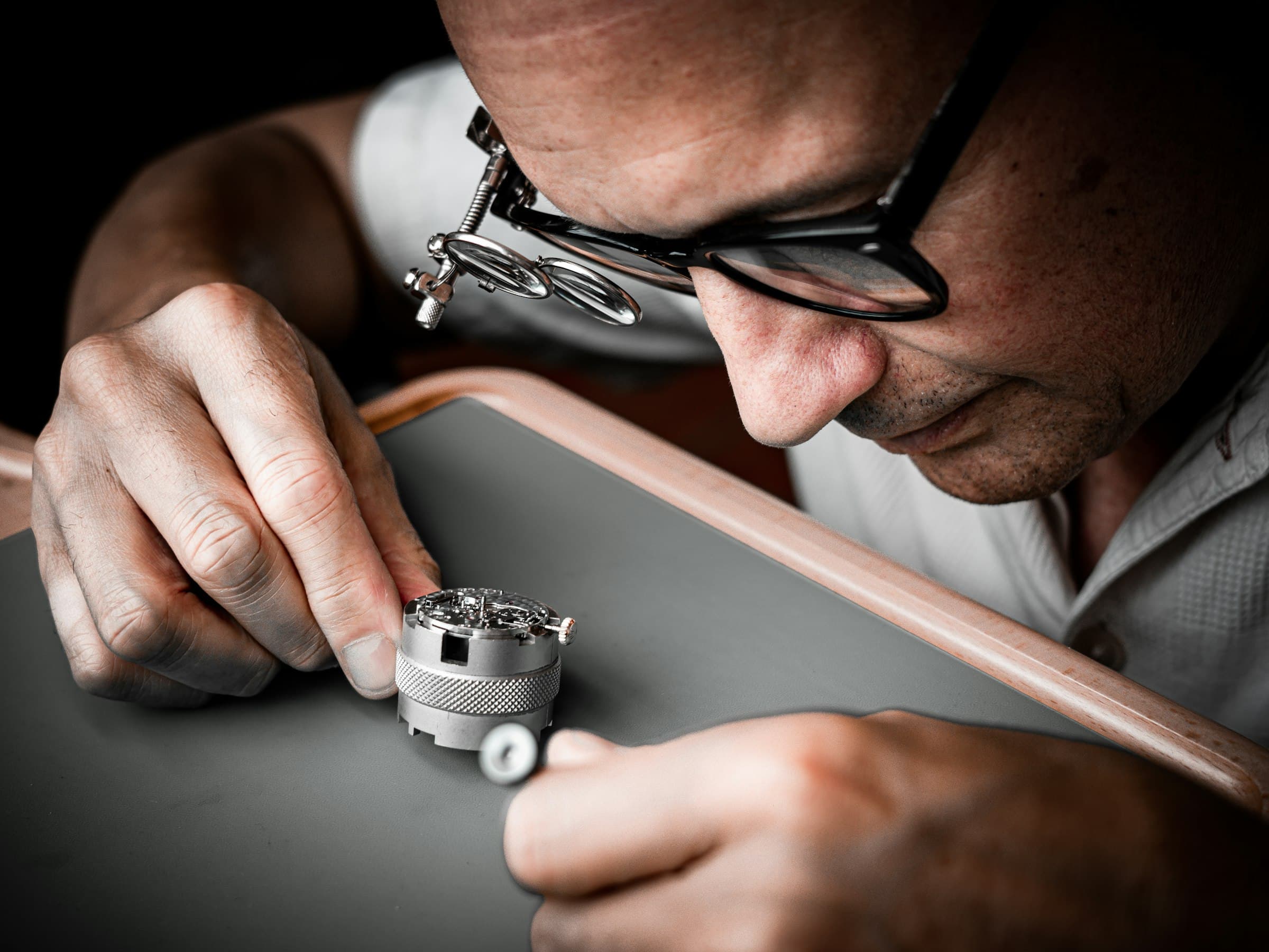Un horloger portant une loupe sur ses lunettes examine un mouvement maintenu dans un support, à son établi.