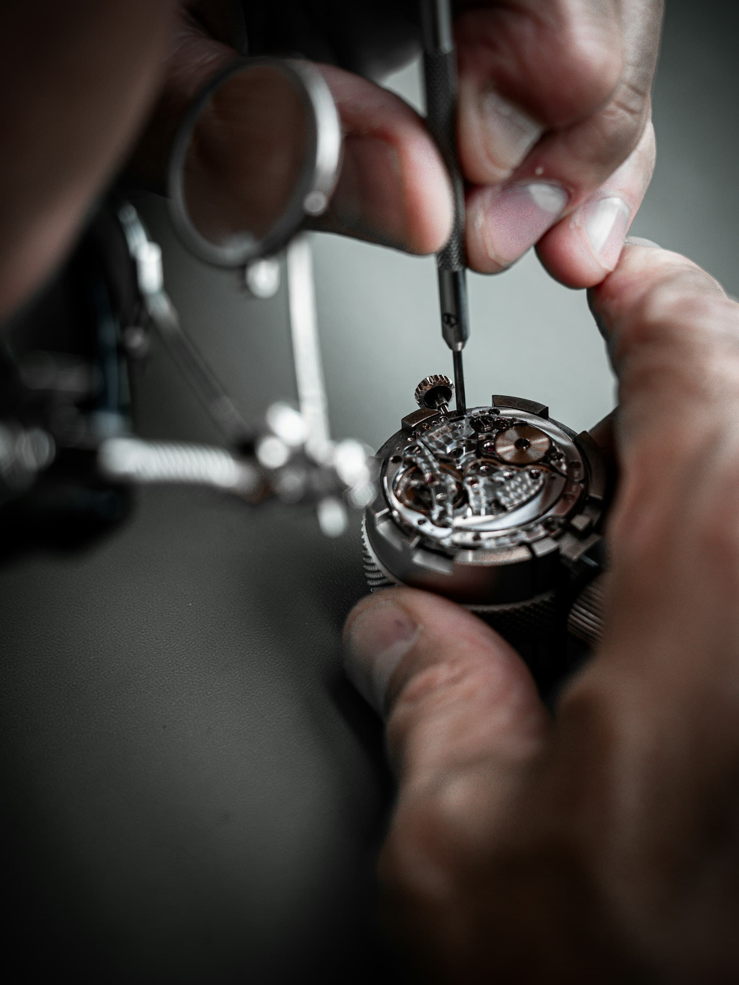 Les mains d’un horloger serrent une vis du mouvement avec un tournevis de précision, sous une loupe articulée.